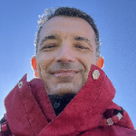 Sebastian Heinz headshot outside with red jacket on.