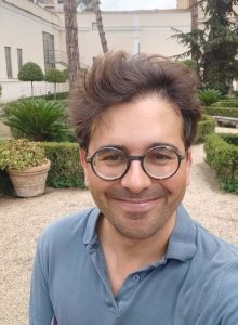 Nicholas Stone smiles in a selfie standing outside in a garden. He wears a blue polo shirt and glasses.