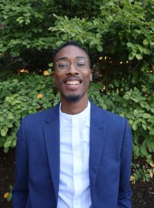 Malcolm Davis smiles outside in front of a green tree wearing a blue dress shirt.