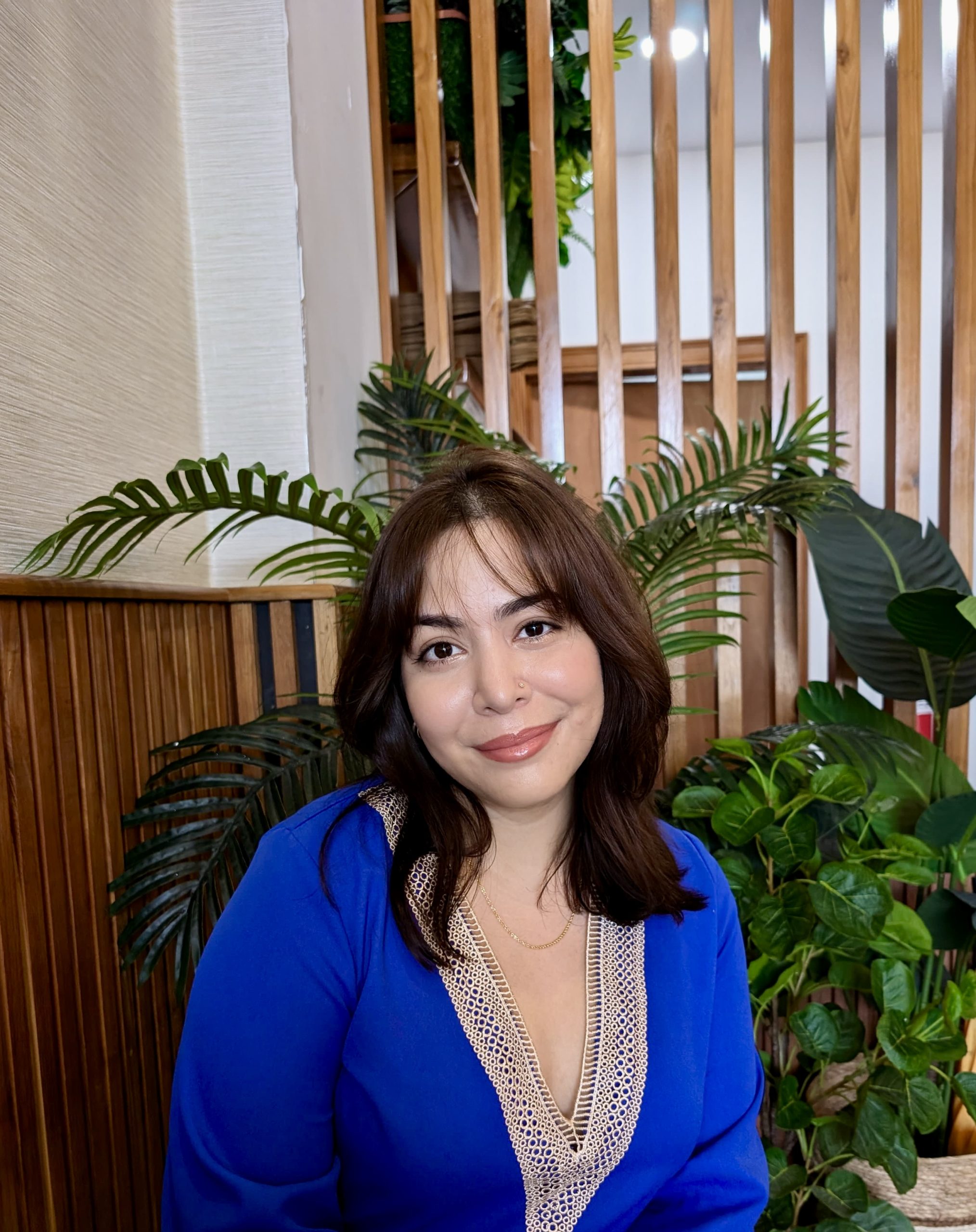Kaylan-Marie sits indoors in front of a plant, smiling and looking at the camera in a blue top。