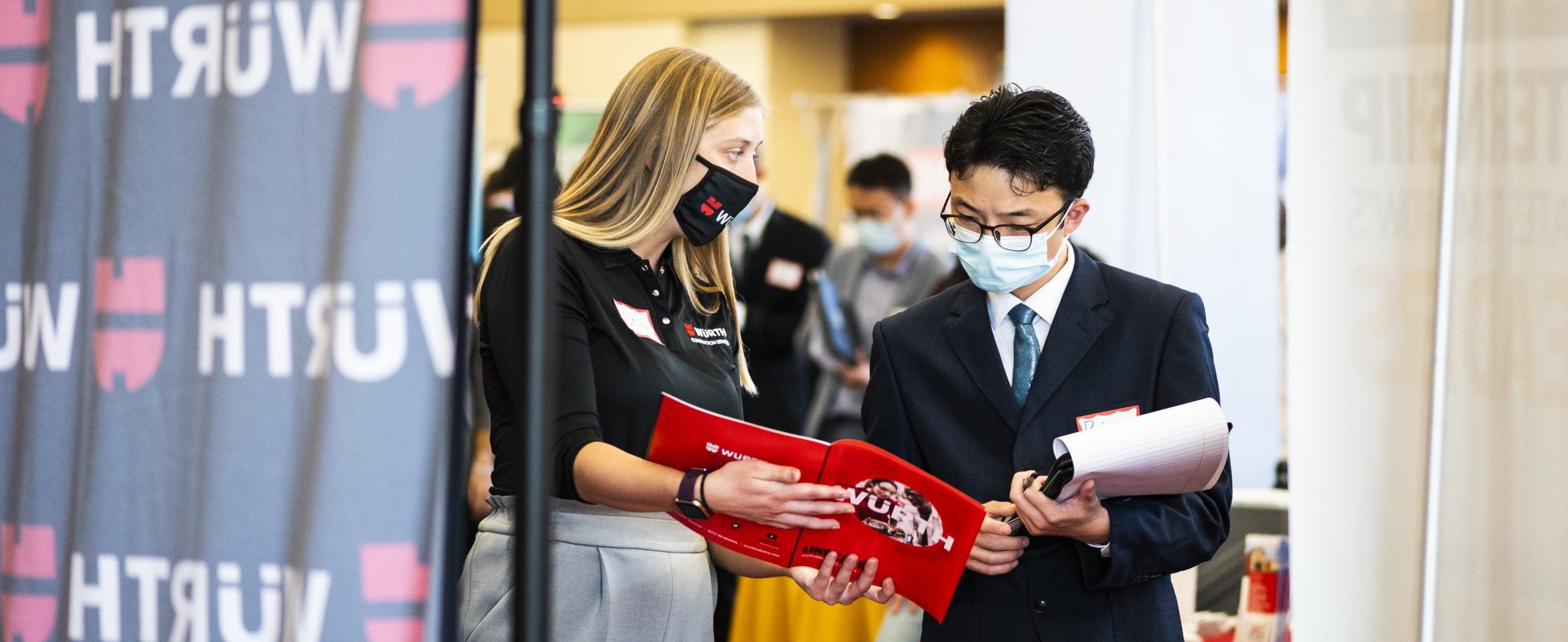 Student and recruiter, wearing masks, speak at the Fall Career and Internship Fair while looking over a packet.