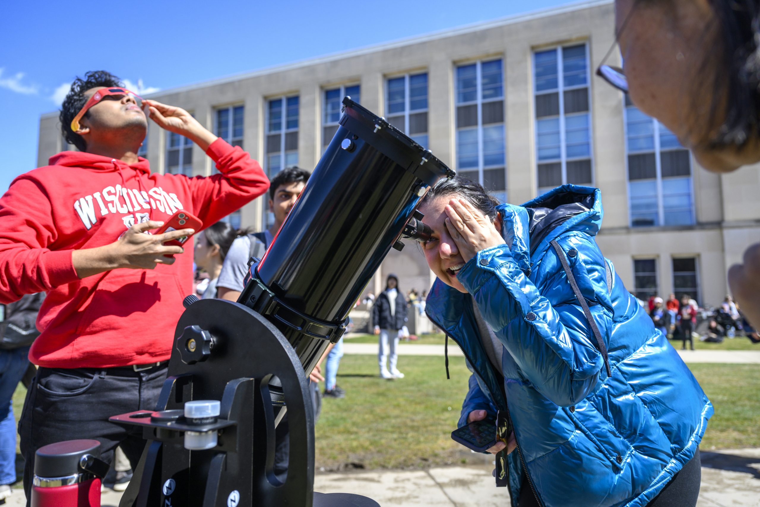At center, a student looks through a telescope while at left, another looks at the sun using solar eclipse glasses during a solar eclipse.