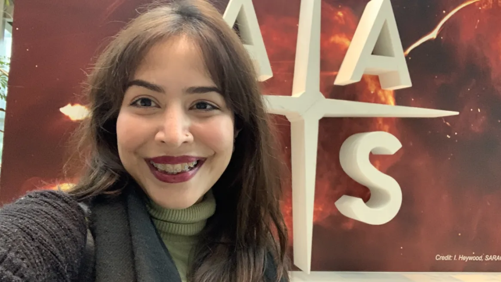 Kaylan Marie Achong smiles in front of the American Astronomical Society sign.