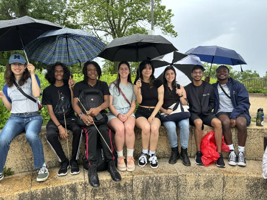 RADIAL students sit outside on a step, holding umbrellas over them.