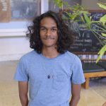 Isaiah Persad poses for a headshot in blue shirt in front of a large green plant.