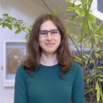 Eliza Canales poses for a headshot in a green shirt in front of a large green plant.